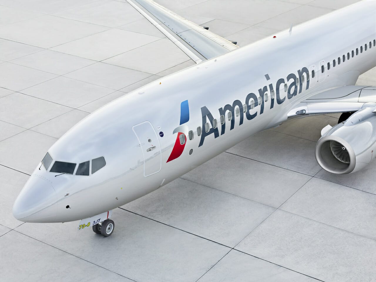 Imagem de um avião da American Airlines no aeroporto Imagem de um avião no aeroporto. Ele é branco, tem o logo azul e vermelho da American Airlines e a palavra "American" escrita em preto.