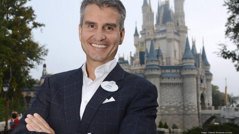 Foto de Josh D'Amaro, presidente do complexo da Disney de Orlando. Ele está sorrindo e o castelo da Cinderela está ao fundo da foto. 