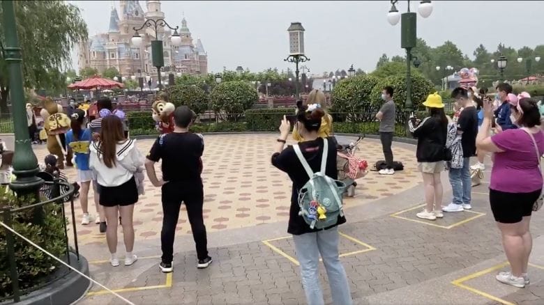 Foto da DIsney de Shanghai mostrando os visitantes praticando o distanciamento devido à pandemia, com marcações no chão onde devem ficar. 
